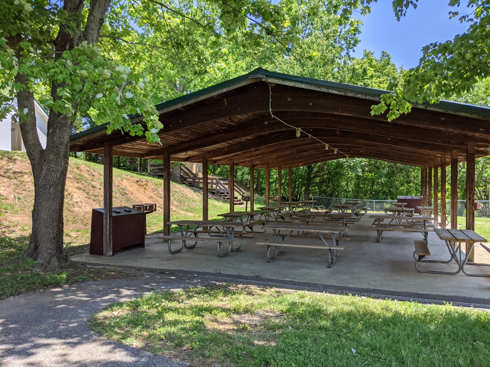 The picnic shelter at Mid County Park is located under shade trees next to the restroom on a sunny summer day.