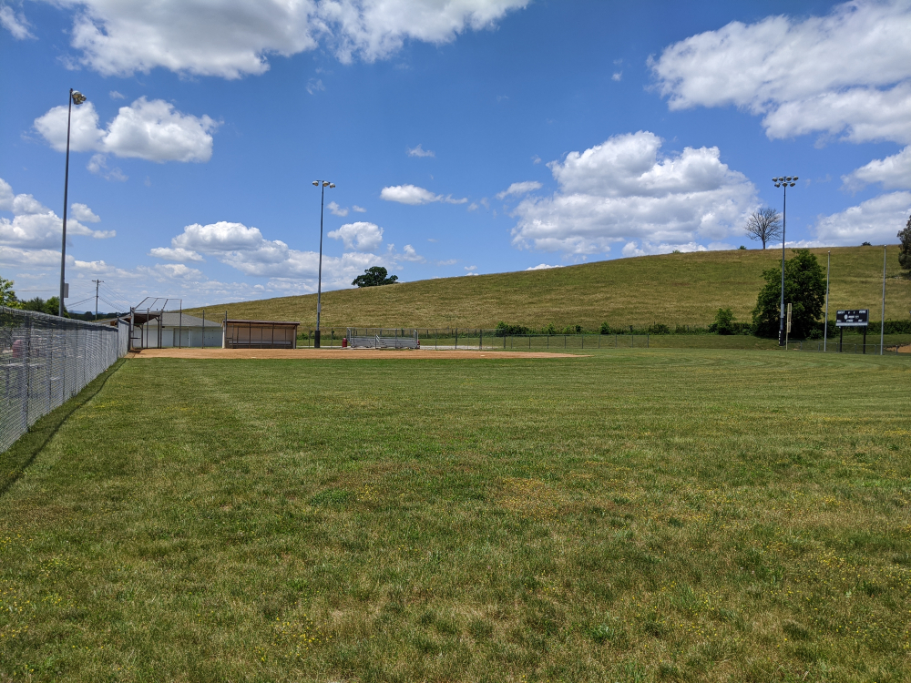 The baseball field at Motor Mile Park is situated under a large grassy hill on a sunny summer day.