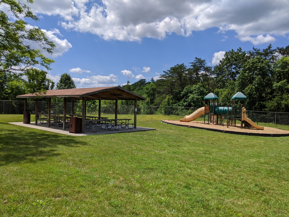The playground and picnic shelter at Plum Creek Park are located next to each other and bordered by trees and green space on a sunny summer day.