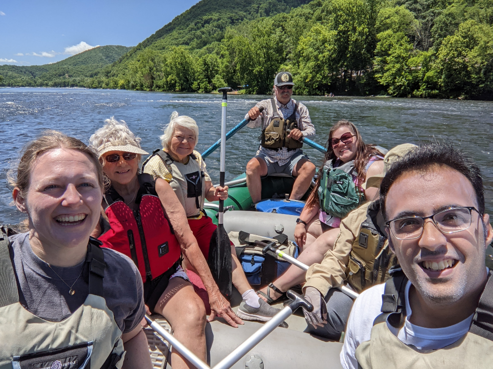 Participants enjoy a raft trip on the New River on a sunny summer day with mountains in the background.